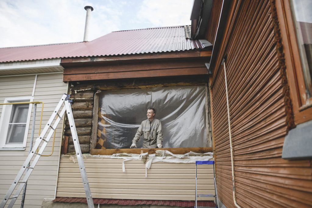 Are Low Efficiency Ratings on Your Windows Costing You? 4 A man peering outside of his home from a large space on the home's siding where a window will be installed.