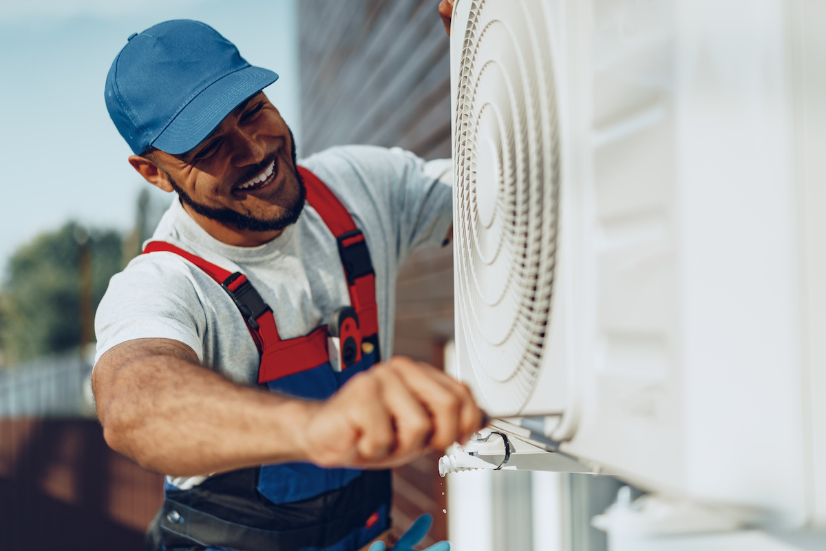 Man wearing a blue hat and overalls while installing a new AC unit and smiling.