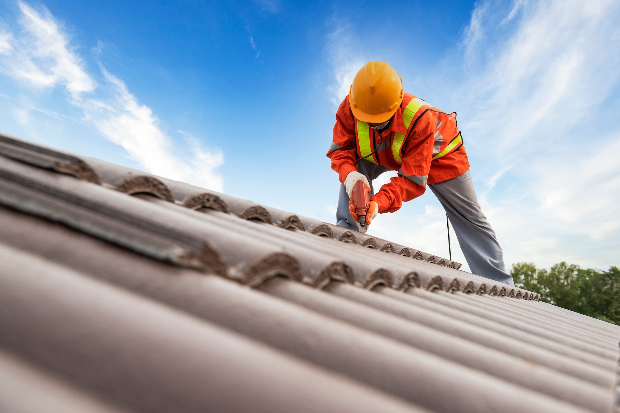 Man in orange safety gear and a hard hat using a drill to repair a roof.