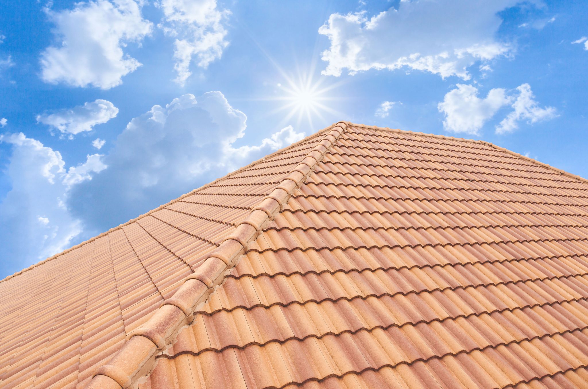Upward view of a terracotta-tiled roof against a bright blue sky with fluffy white clouds and the sun shining brightly.
