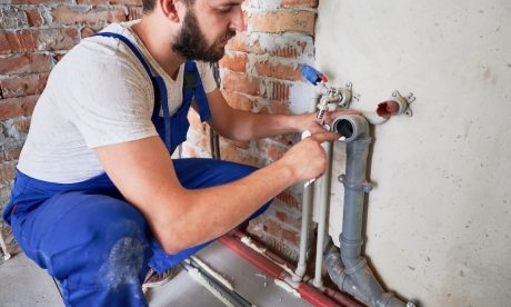 Main Home 21 A plumber wearing a white hard hat and blue overalls works on a sewer pipe attached to a brick wall in an unfinished building.