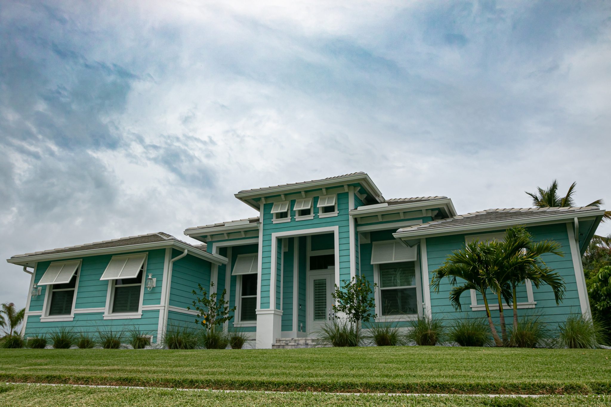 A single-story turquoise house with white trim, large front entrance, shuttered windows, and a well-maintained lawn under a cloudy sky.